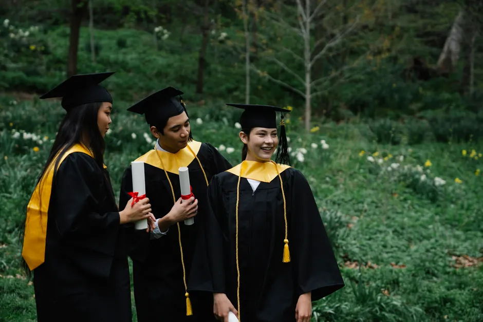 Three graduates in gowns and caps celebrating outdoors among greenery.