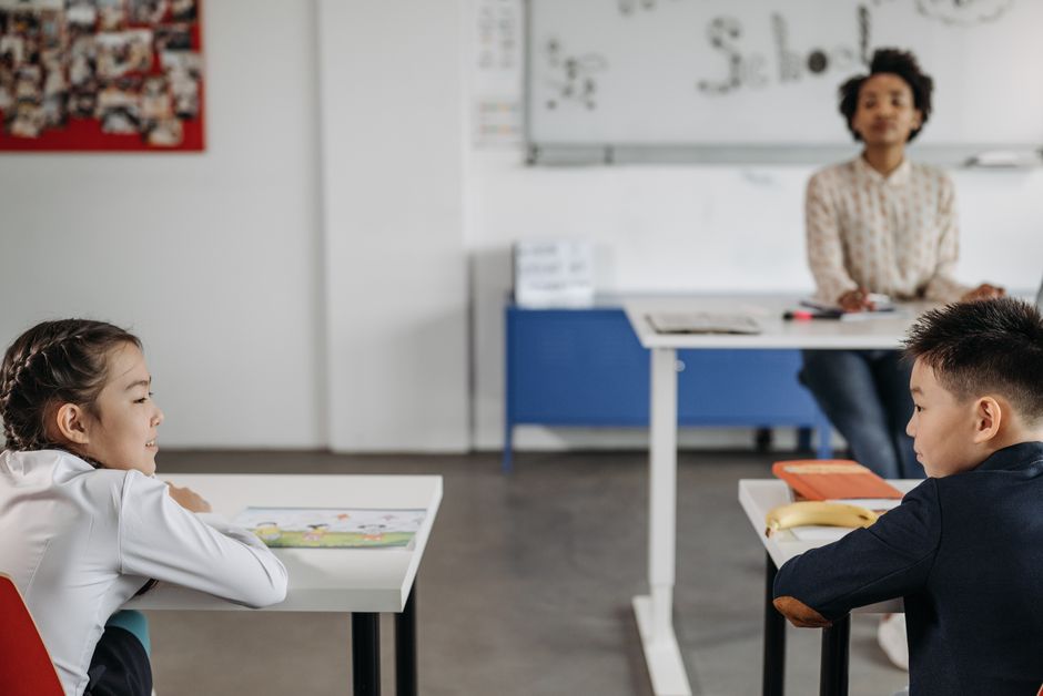 Two students engage in a classroom setting with their teacher in the background.
