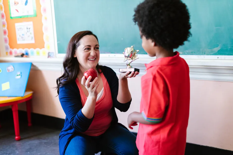 A teacher holding an apple and a student holding a trophy in a classroom setting.