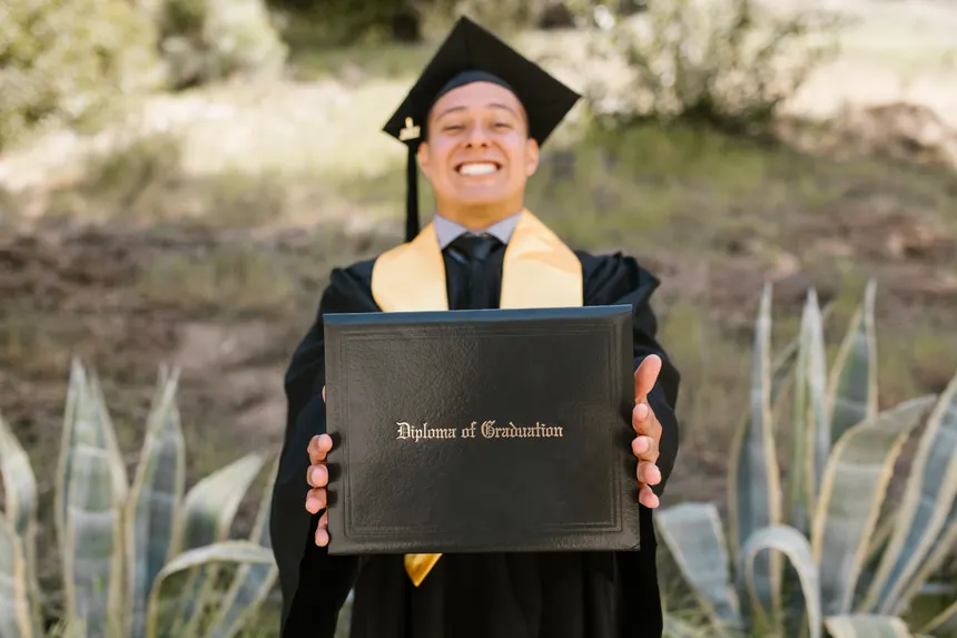 Smiling graduate in academic dress displaying diploma outdoors, symbolizing achievement.