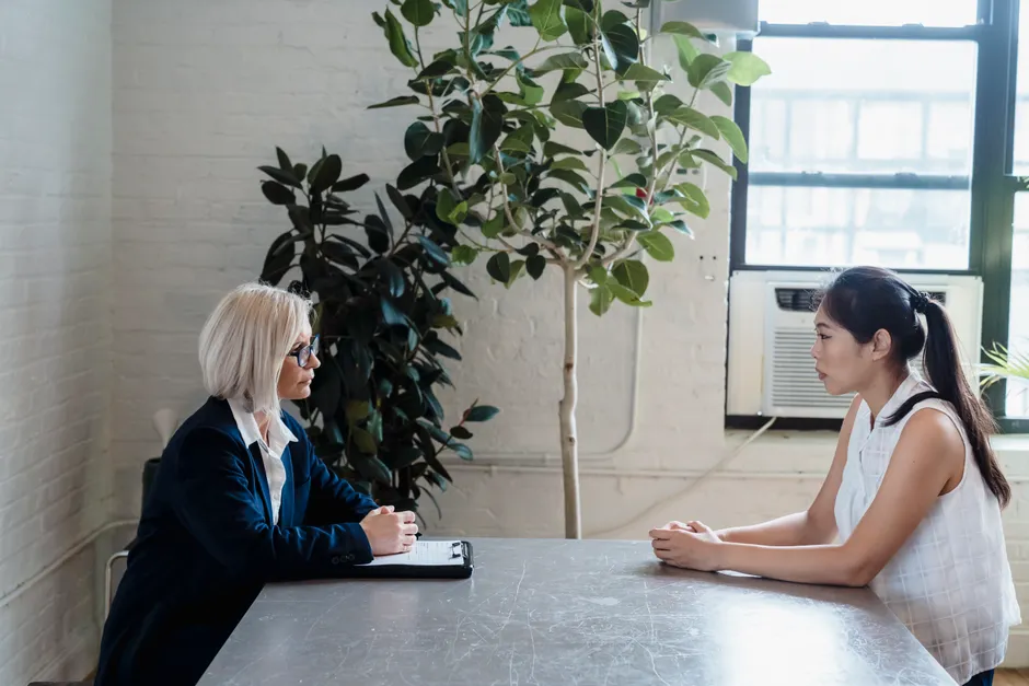 Two women sit across from each other indoors in a serious conversation setting.