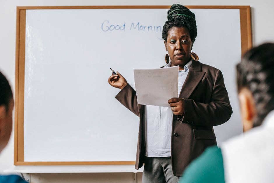 Concentrated adult African American female teacher wearing formal suit standing near whiteboard and talking to pupils in light classroom