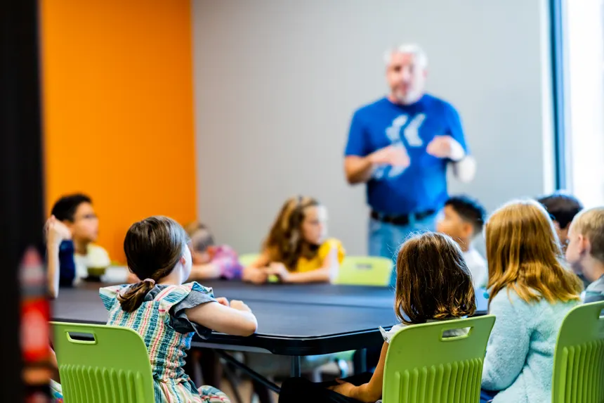 Children attentively listen to a teacher during a group activity in a colorful classroom setting.
