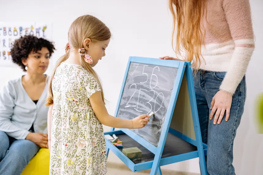 A young girl writes numbers on a chalkboard in a classroom, supervised by adults.