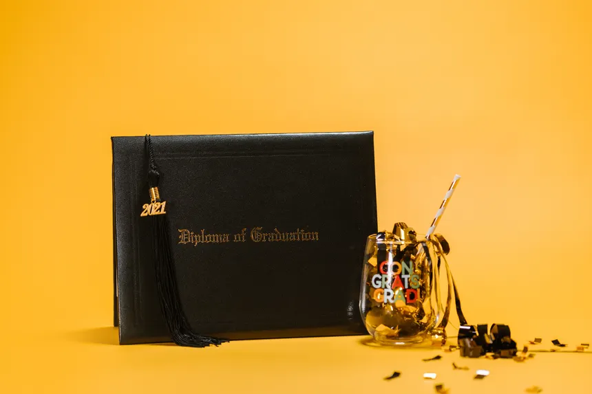 Black diploma and glass with confetti against a yellow background, symbolizing graduation celebration.